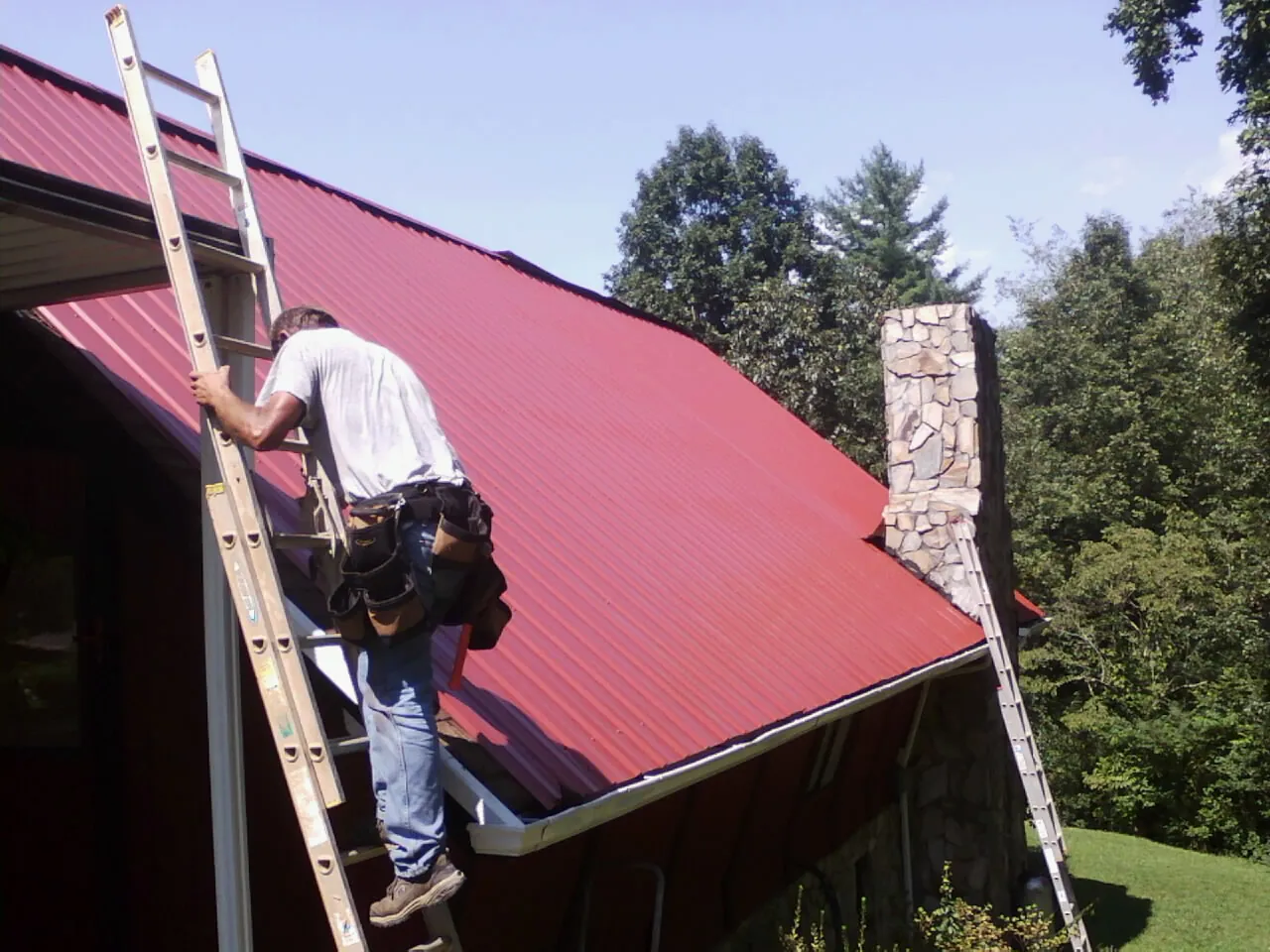 Roofer on a ladder beside a red metal roof and stone chimney — Soffit & Fascia Repair by Roofing Brothers in Muscatine